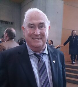 Man, white hair, suit and tie, name is John Simpson