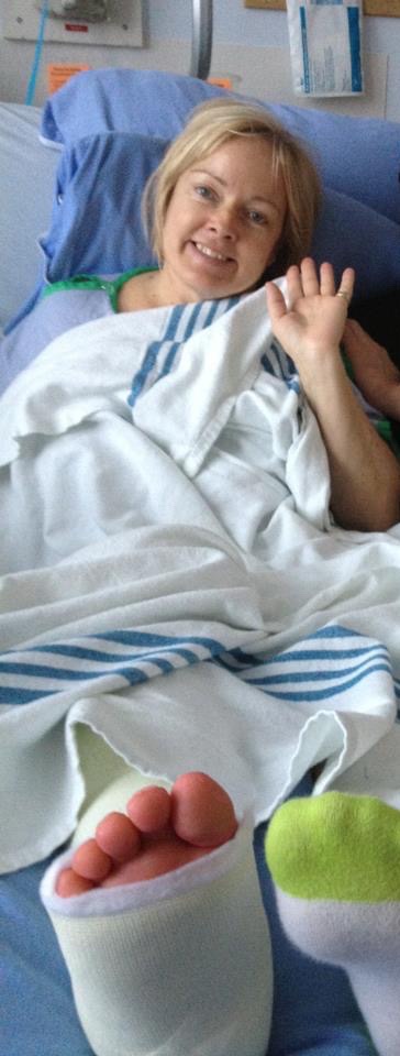 Debra, a lady with blonde hair, lays in a hospital bed with her foot in a brace. She waves to the camera.